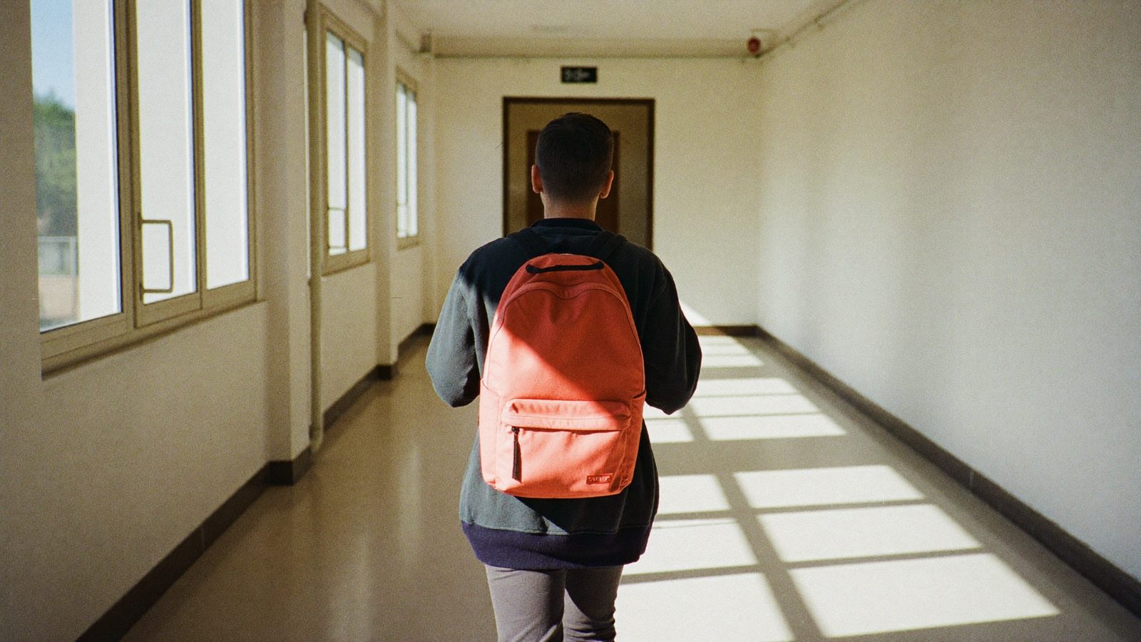 Studente di spalle che percorre il corridoio deserto di una scuola superiore con lo zaino sulle spalle