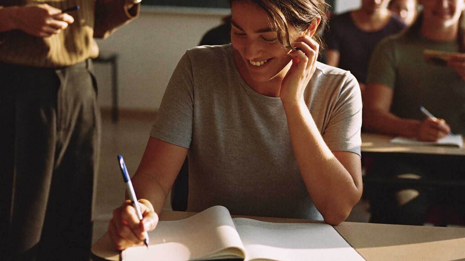 Piccolo gruppo di adulti di provenienze diverse seduti ai banchi di un'aula serale, uno di loro scrive su un quaderno mentre un docente spiega, luce calda di fine giornata