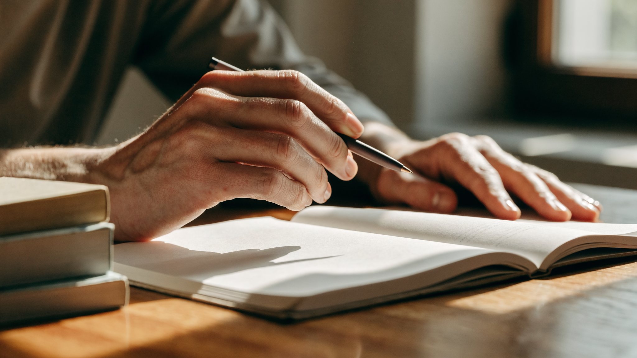 Le mani di un adulto al tavolo di casa, con una penna sopra un quaderno aperto e alcuni libri di testo accanto.