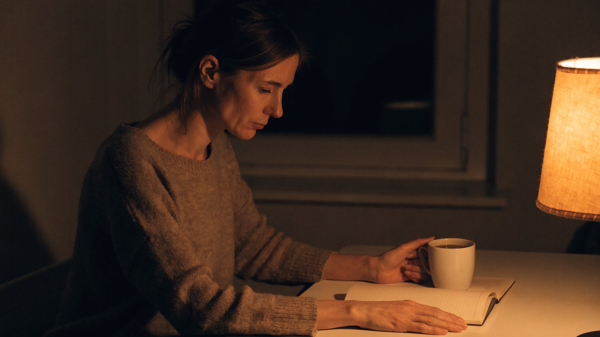 Una donna studia la sera al tavolo di casa alla luce di una lampada, con un quaderno aperto e una tazza accanto.
