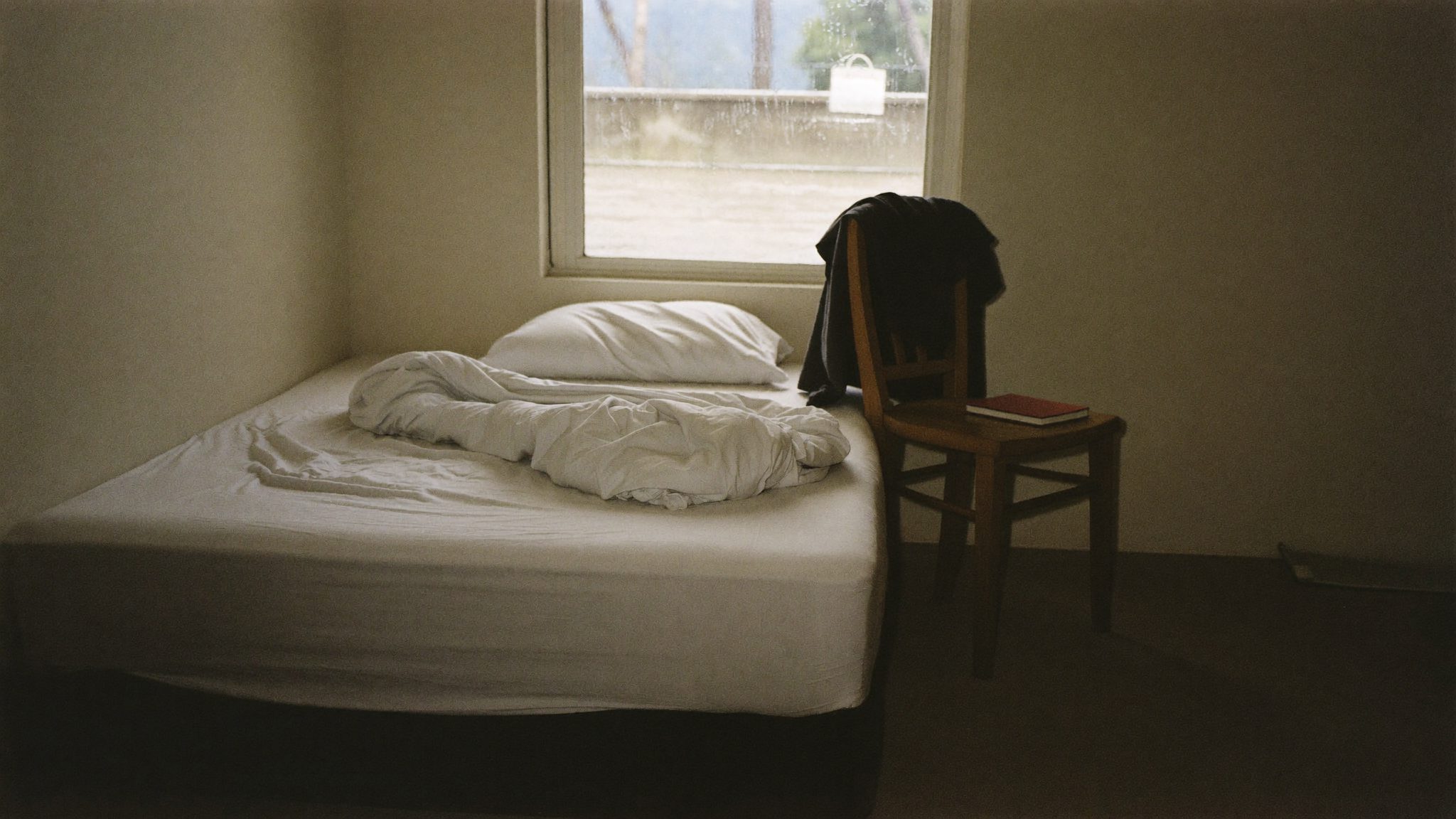 Camera da letto adolescenziale vuota con il letto sfatto e un quaderno chiuso sul pavimento, nella luce grigia di un giorno di pioggia