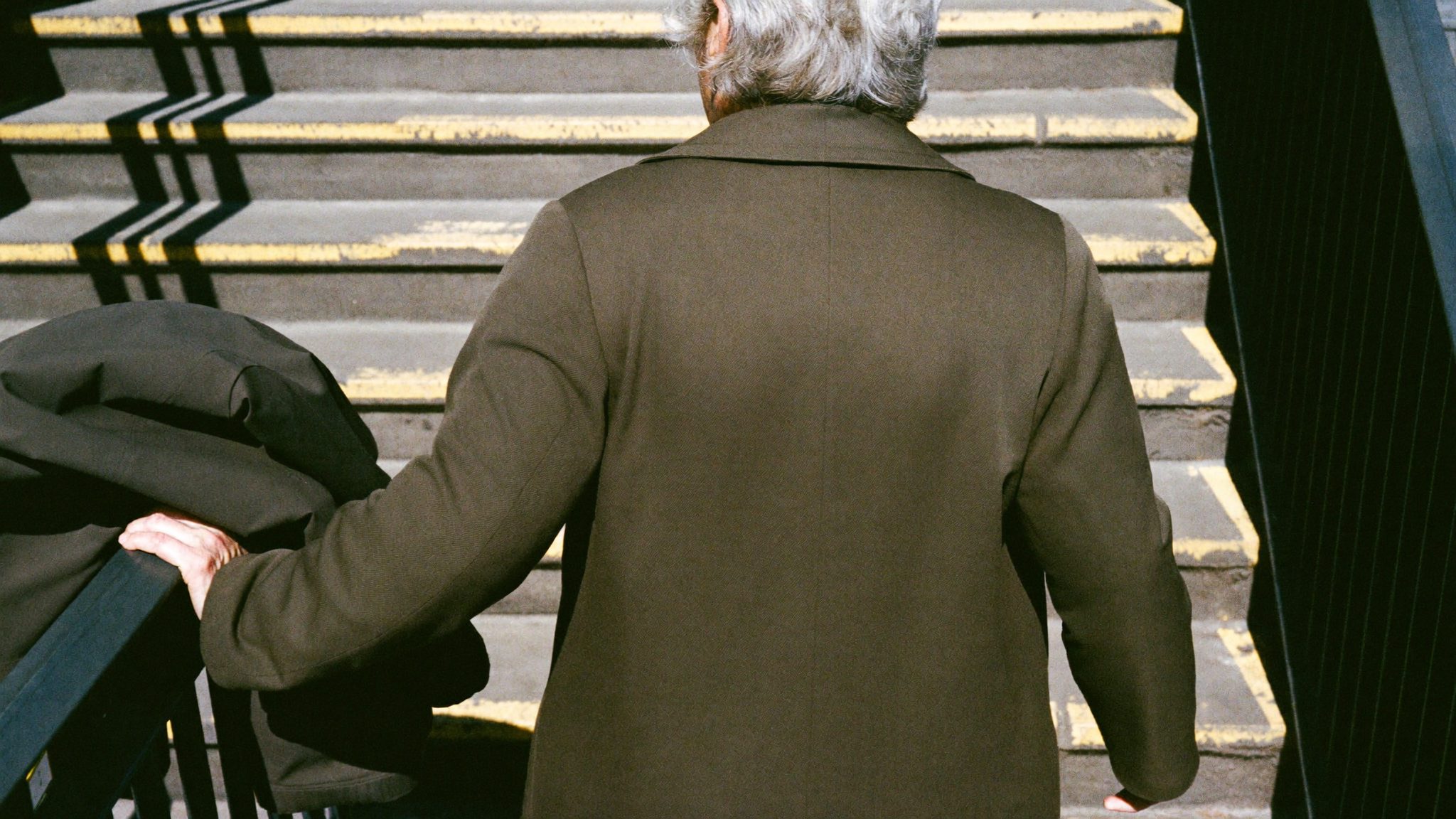 Donna anziana di spalle con cappotto scuro sale gli scalini della metro, ombre nette di mezzogiorno sui gradini gialli