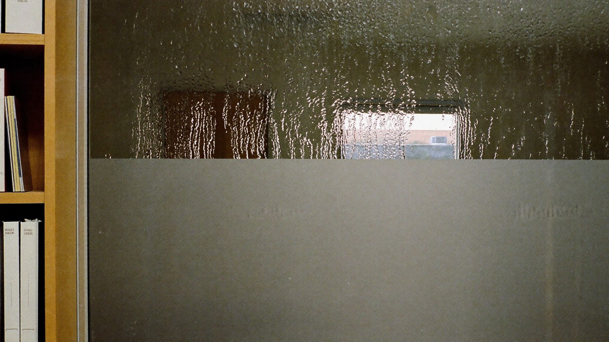 Finestra bagnata dalla pioggia in una stanza istituzionale vuota, gocce d'acqua sul vetro opaco, scaffale con raccoglitori sul lato sinistro