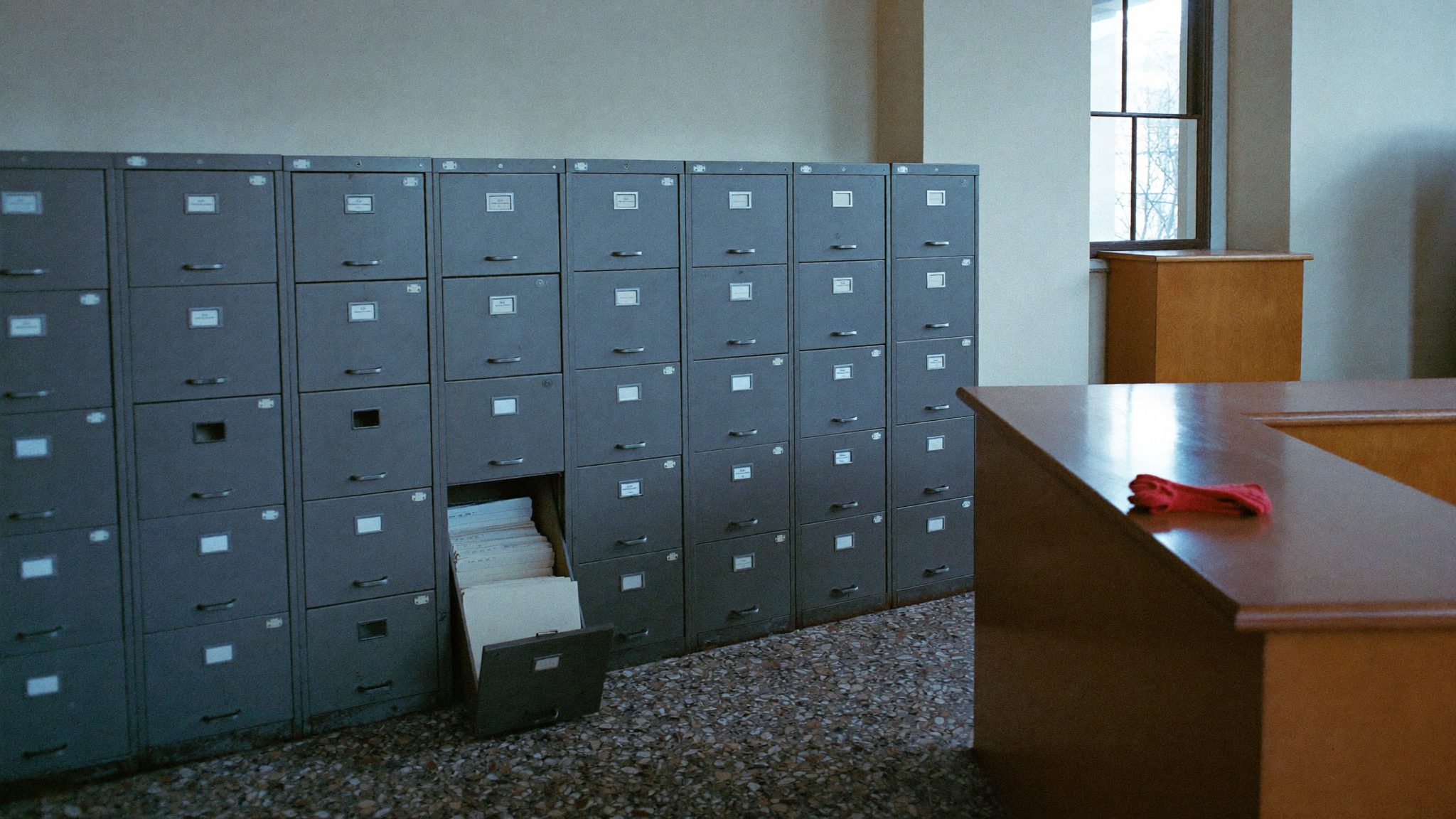 L'archivio vuoto di una segreteria al mattino presto, con un cassetto aperto pieno di cartelline e un paio di guanti corallo sul bancone