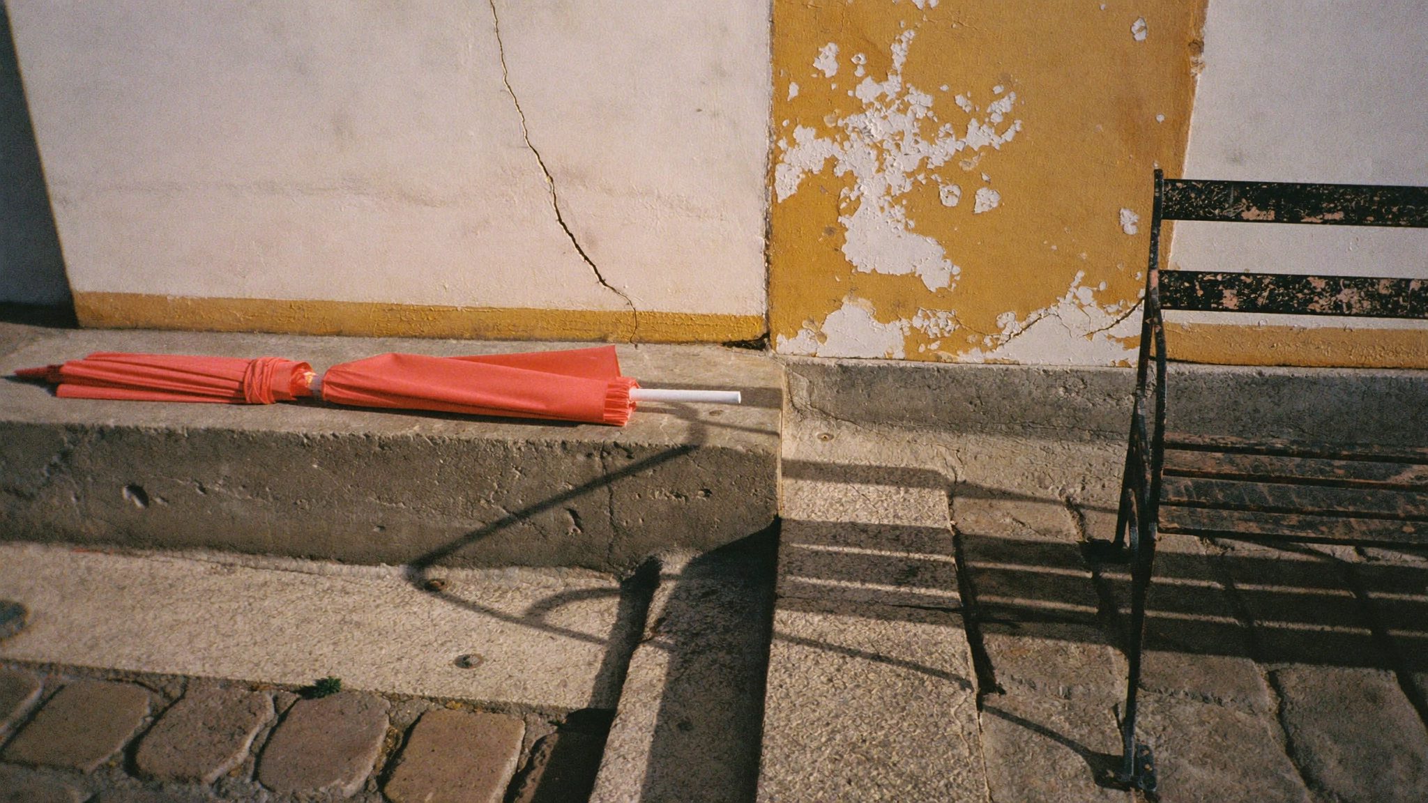 Piccola piazza italiana con ombrello corallo appoggiato al muro e panchina in ghisa con vernice scrostata, luce del tardo pomeriggio