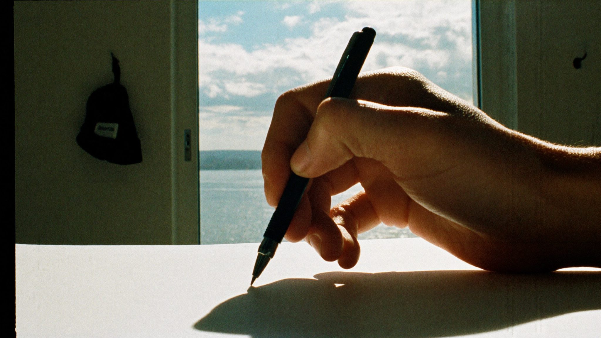 Mani che tengono una penna su un foglio di carta davanti a una finestra luminosa, con un cappello corallo sfocato sullo sfondo