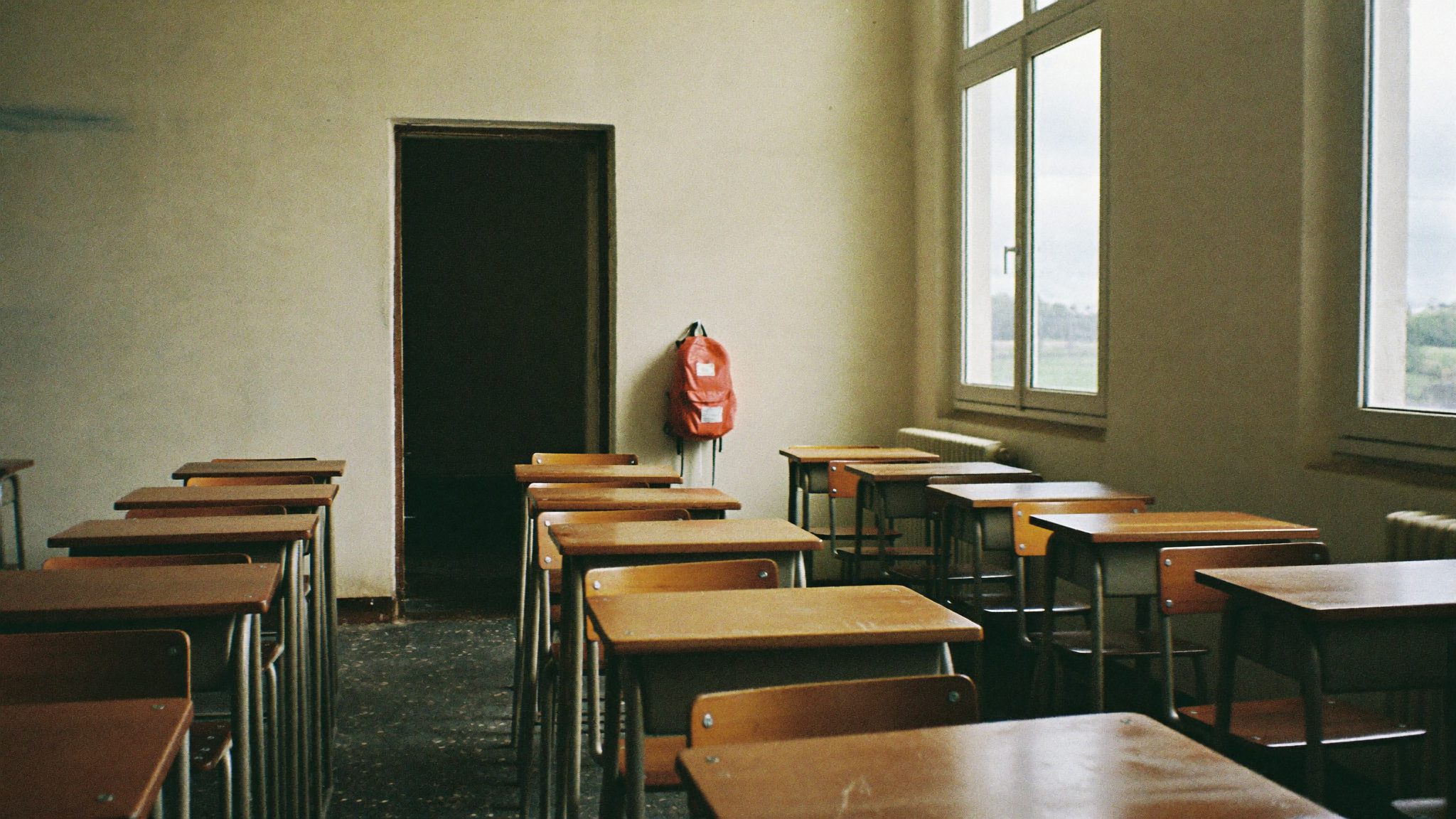 Aula scolastica vuota con file di banchi in legno consumato e uno zaino color corallo appeso a una sedia vicino alla porta