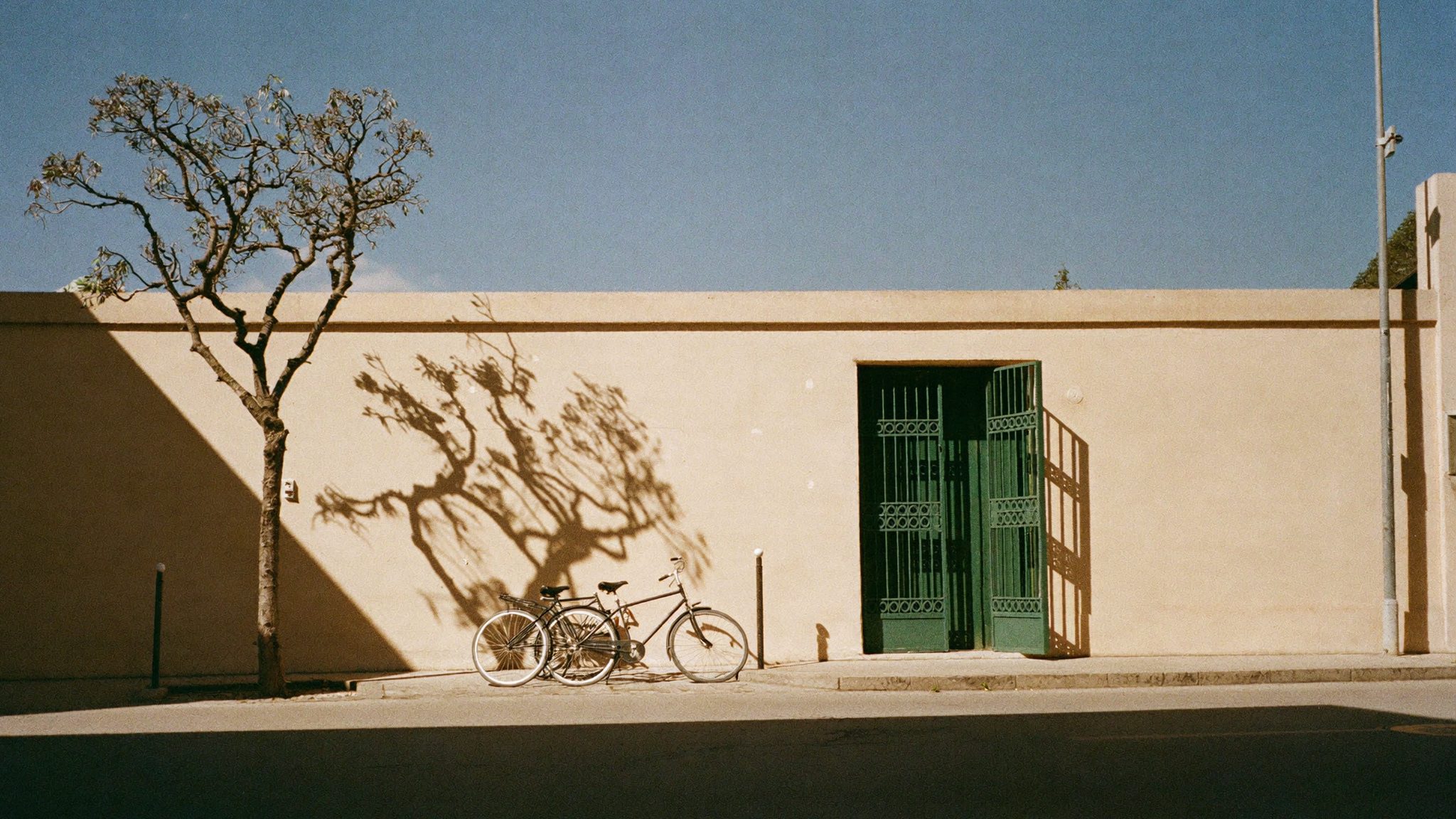Un muro color ocra con un cancello verde aperto e due biciclette appoggiate, sotto il sole di mezzogiorno