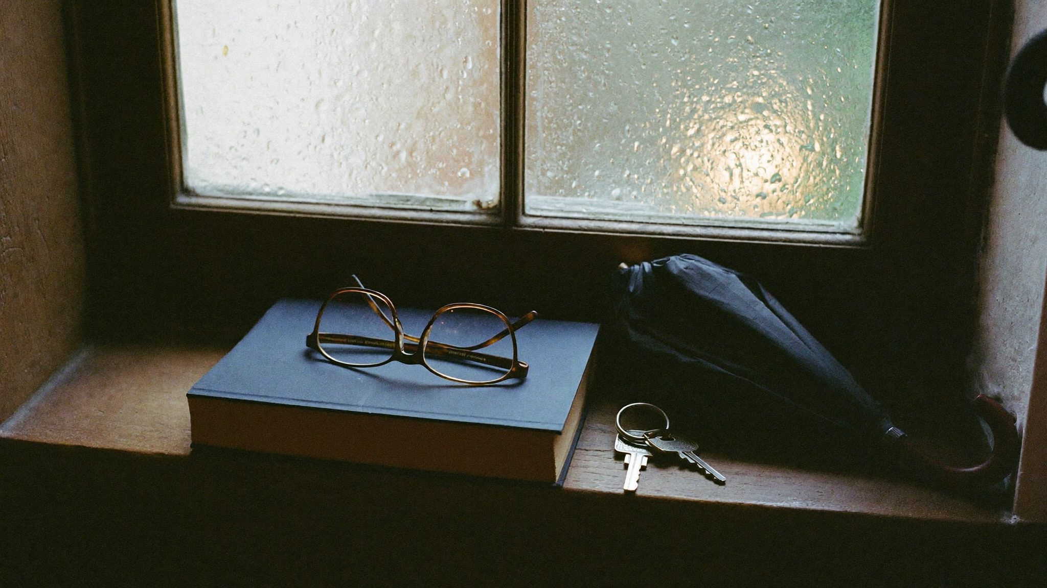 Su un davanzale in un giorno di pioggia: un libro chiuso, un paio di occhiali da lettura, le chiavi di casa e un ombrello