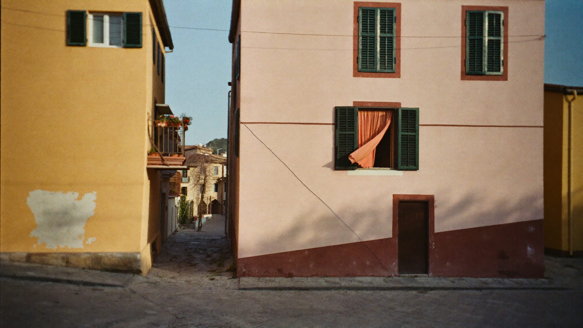 Una via stretta di una cittadina italiana al tramonto, con una tenda corallo che esce da una finestra aperta