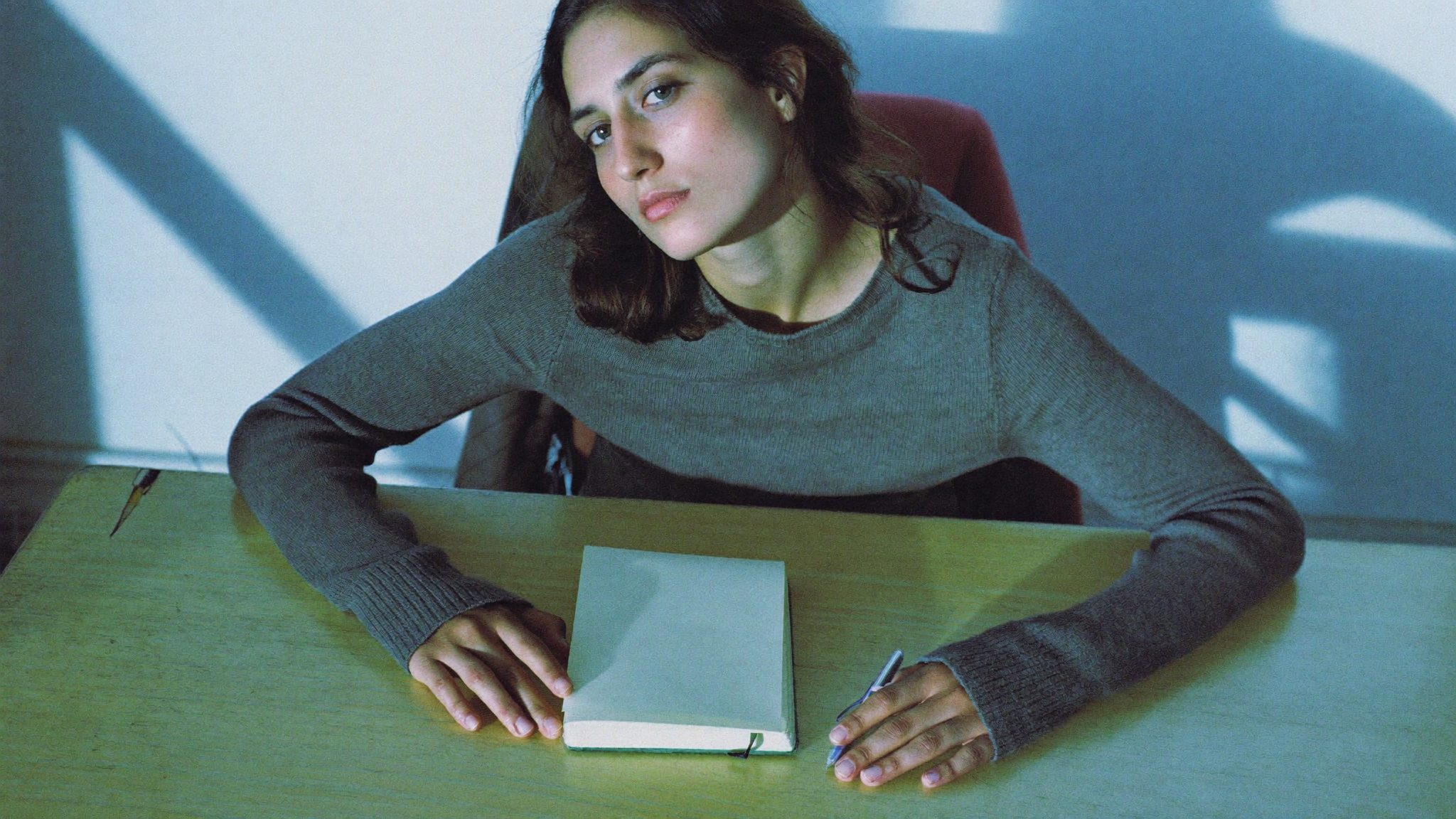 Giovane donna con maglione grigio al tavolo da cucina, quaderno chiuso davanti a sé, luce fredda di mattina dalla finestra