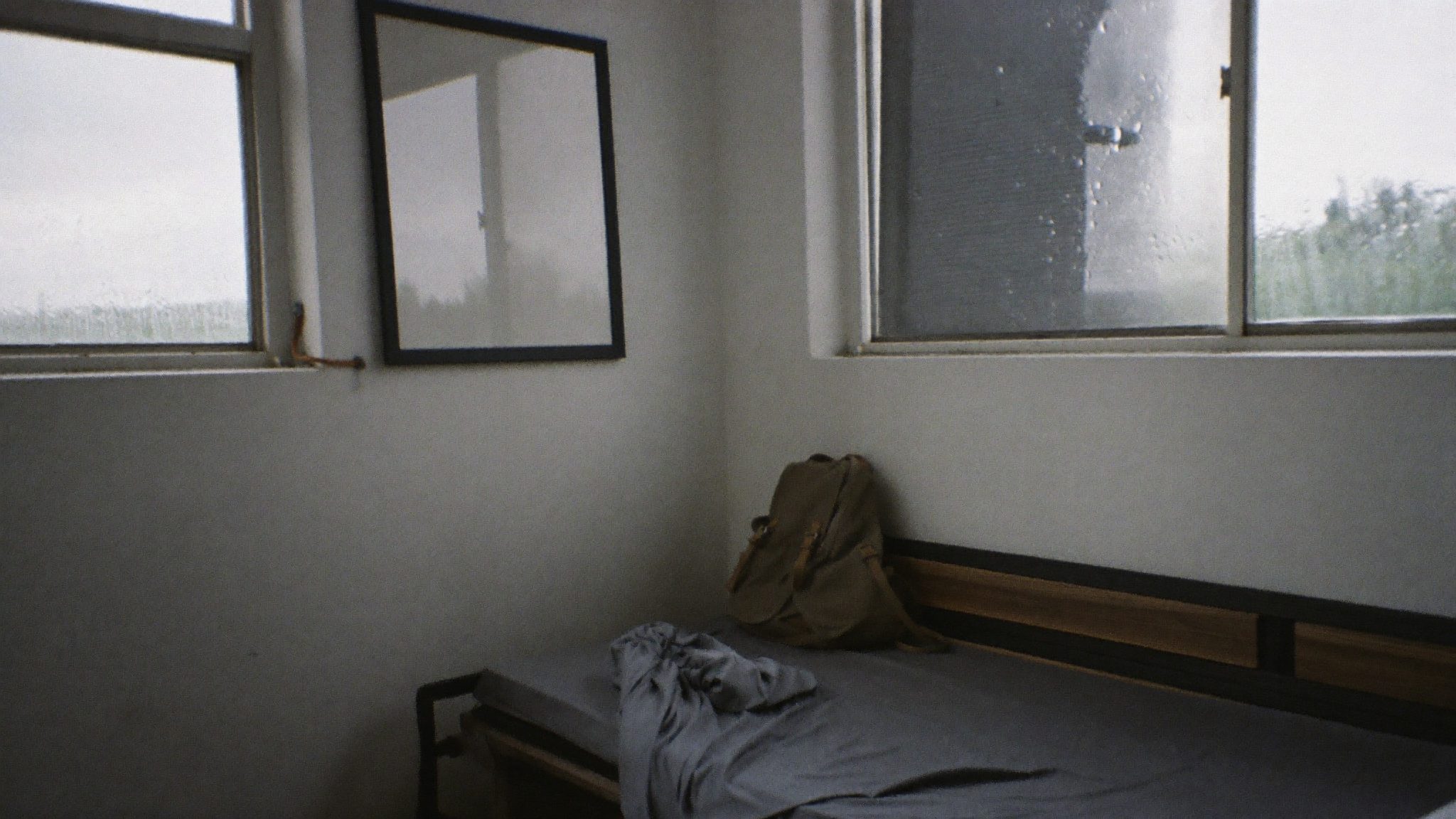 Camera vuota con letto disfatto e zaino abbandonato, finestre con vetri bagnati di pioggia e luce grigia