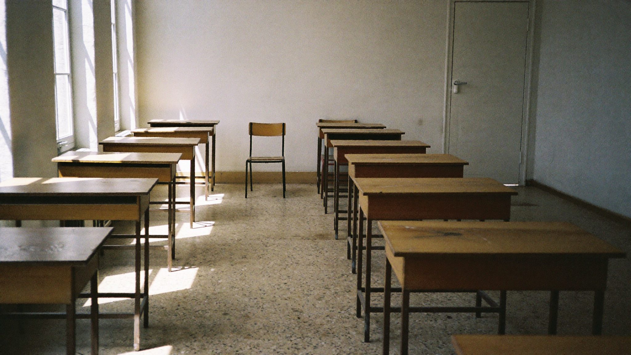 Aula scolastica italiana vuota con banchi di legno graffiati e luce di mezzogiorno che filtra dalle finestre