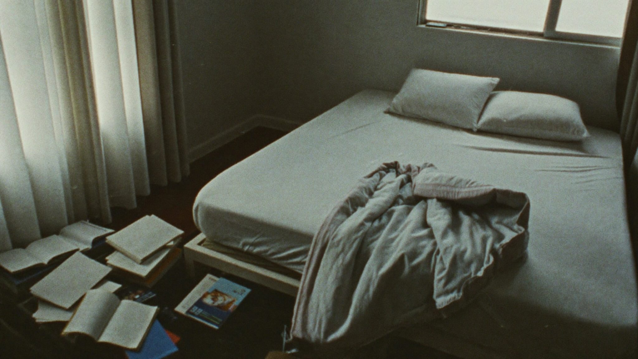 Cameretta di adolescente vuota con il letto disfatto e libri impilati sul pavimento, luce diffusa e piatta di giornata coperta che entra dalla finestra