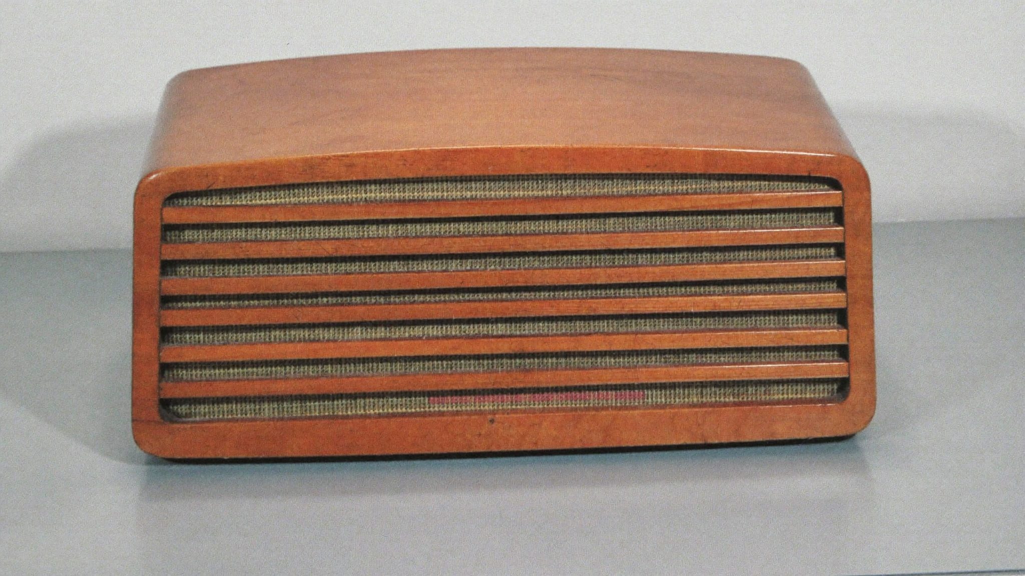 Una vecchia radio di legno con la griglia di stoffa attraversata da listelli, sola su un fondo chiaro