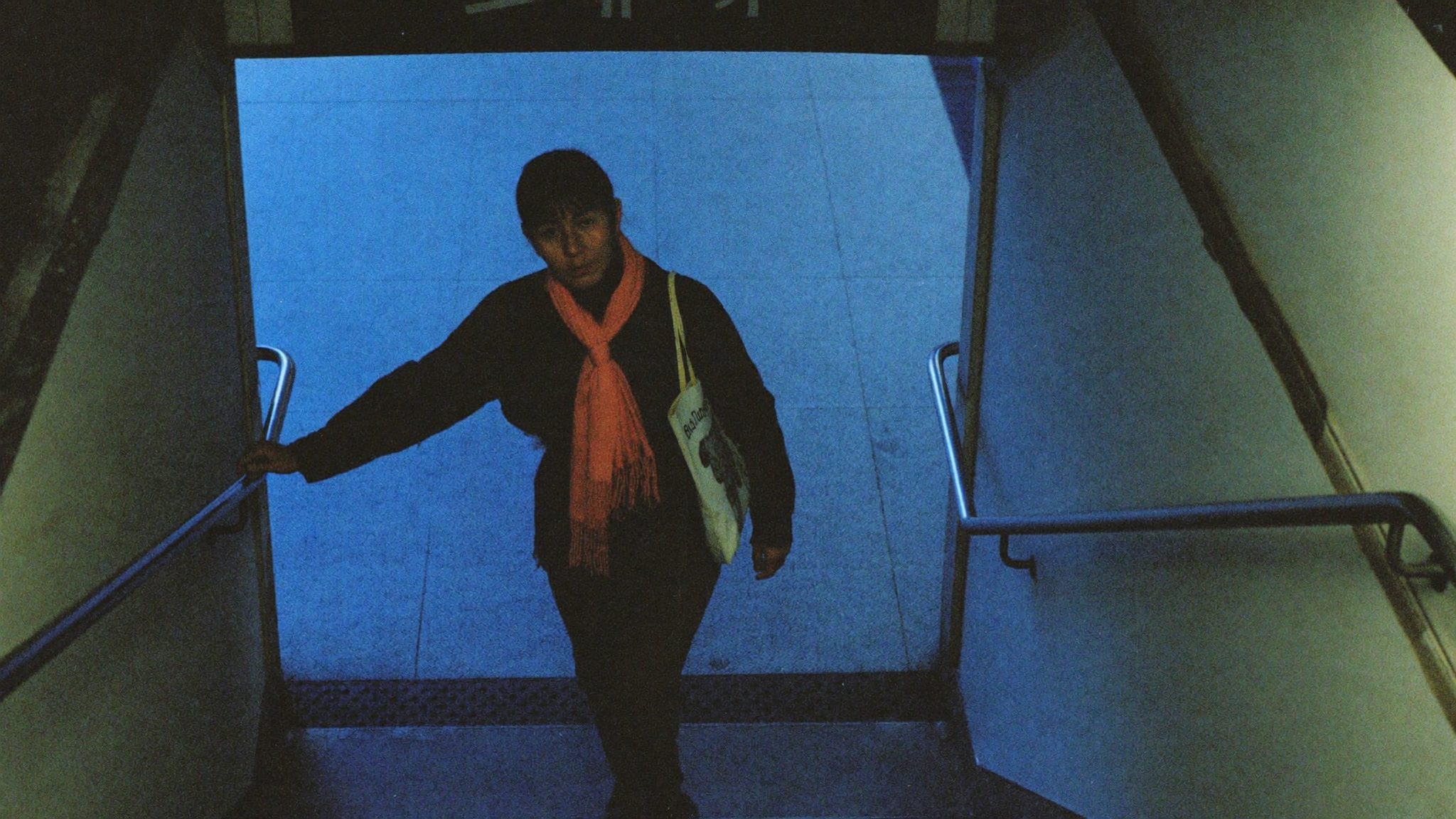Insegnante sulla quarantina che sale le scale della metropolitana con una sciarpa corallo, luce blu dell'ora di punta, non guarda in camera