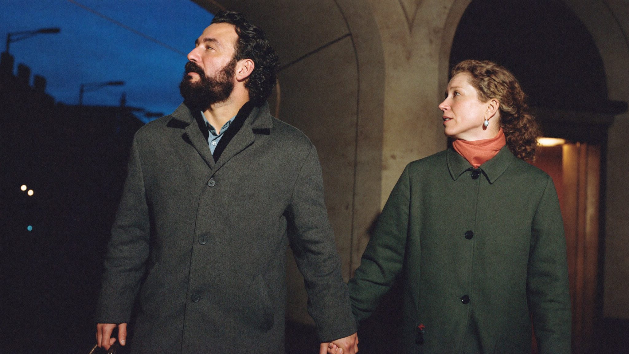 Un uomo con la barba e una donna con una sciarpa corallo camminano mano nella mano sotto un portico all'ora blu