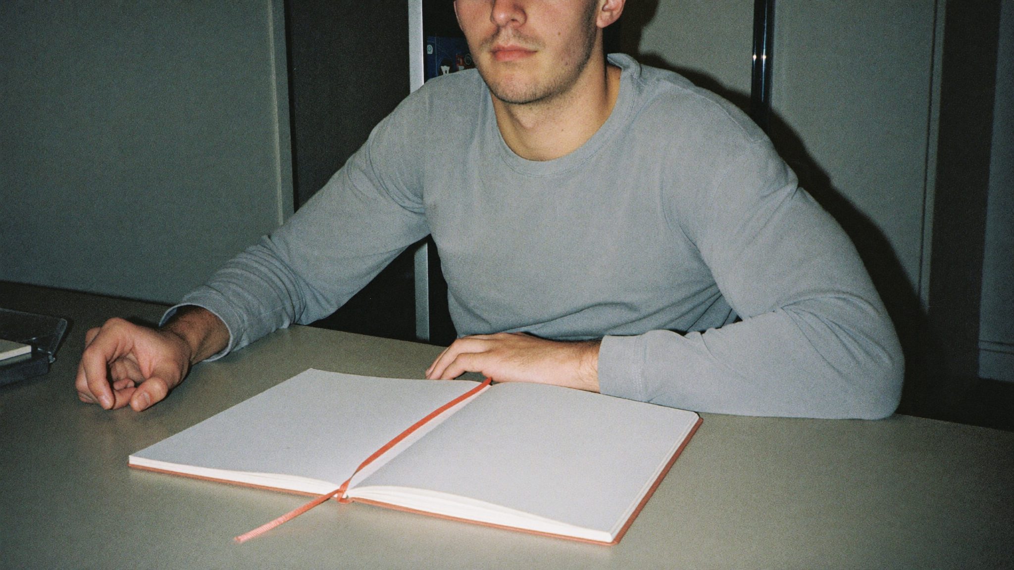 Giovane uomo di vent'anni seduto a un tavolo di cucina con un quaderno chiuso e un segnalibro color corallo visibile sul bordo, luce fredda al neon, sguardo altrove