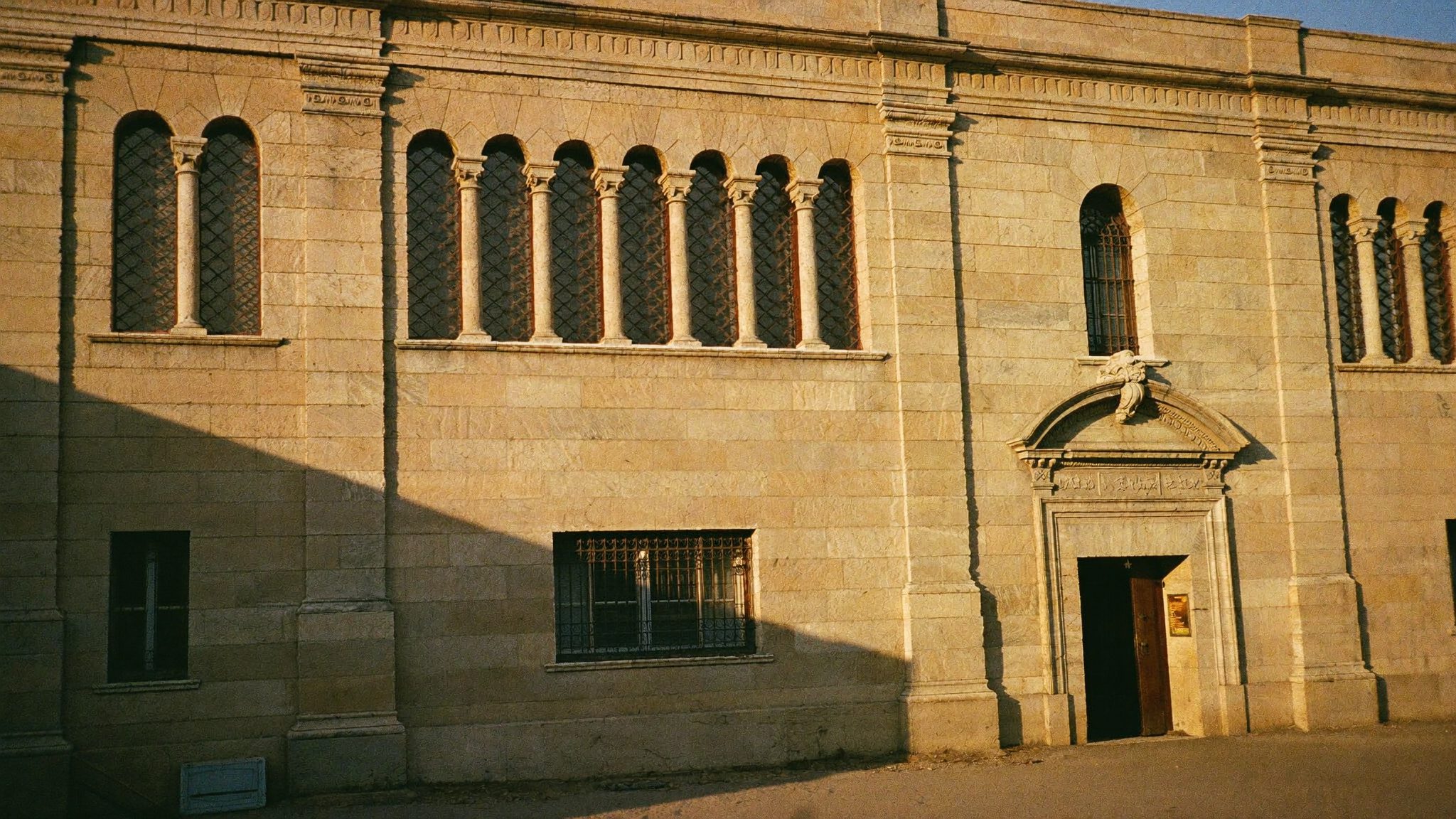 Facciata in pietra di un edificio scolastico italiano degli anni Trenta con finestre ad arco, illuminata dalla luce radente del tardo pomeriggio che proietta lunghe ombre sulla pietra