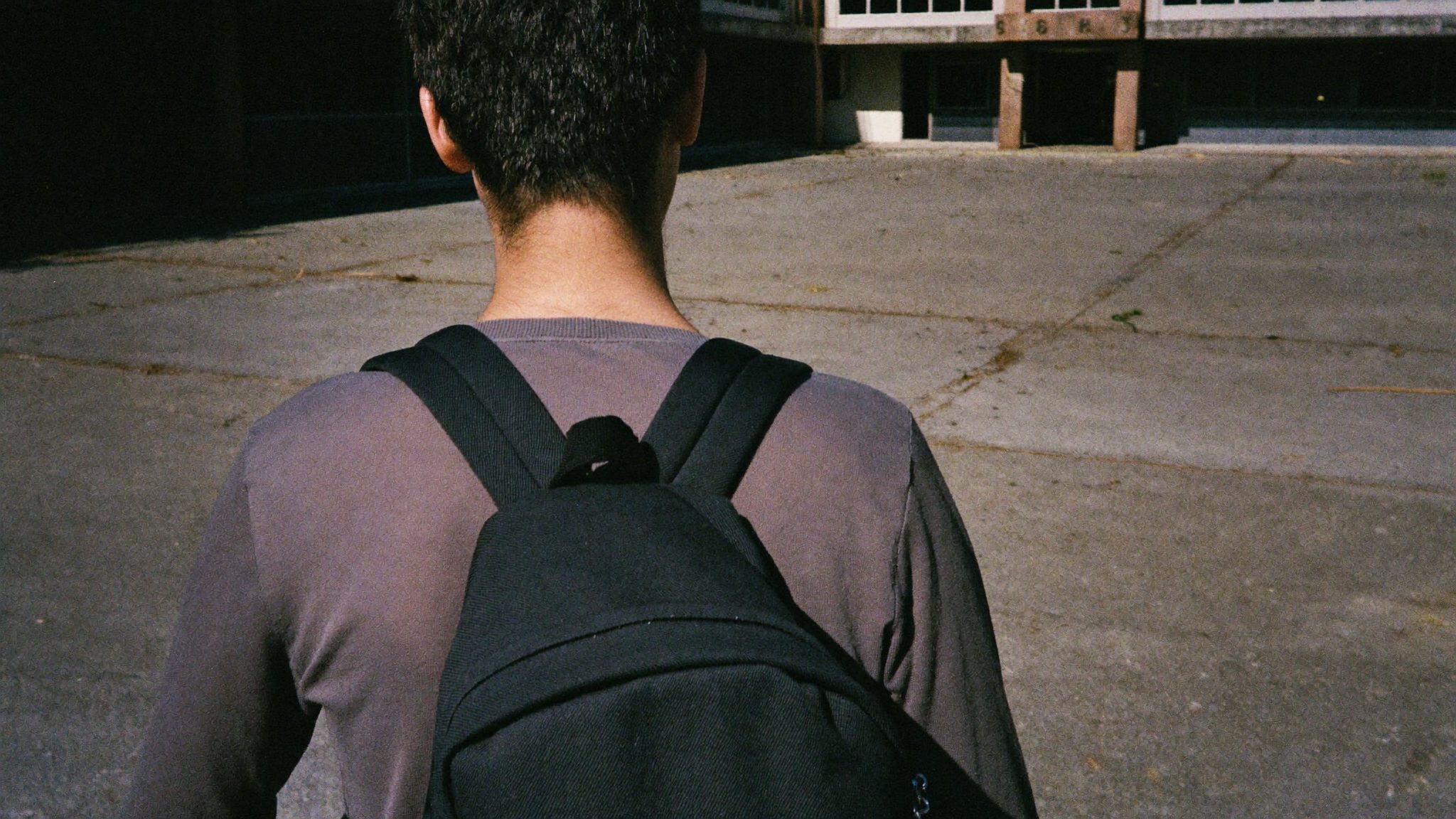 Studente adulto di spalle con zaino scuro cammina verso l'ingresso di un edificio scolastico, ombre nette del sole di mezzogiorno sul cemento