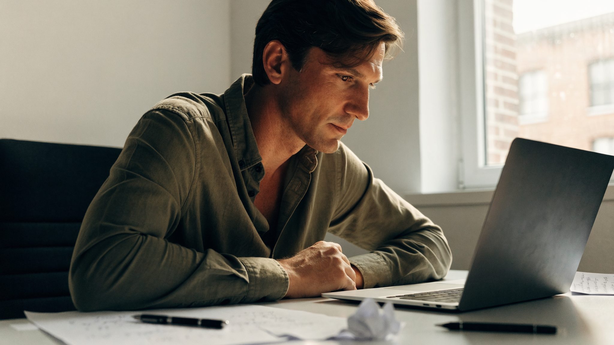 Uomo di circa 35 anni seduto a una scrivania ordinata, con un laptop aperto e uno smartphone accanto, luce naturale da una finestra laterale, espressione serena e concentrata, nessun testo leggibile sugli schermi