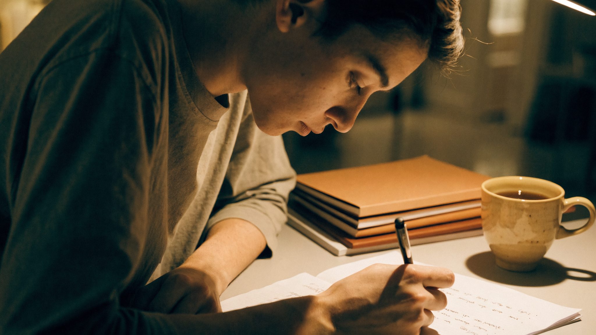 Un ragazzo studia al tavolo di casa con più quaderni aperti e una lista scritta a mano accanto, alla luce di una lampada.